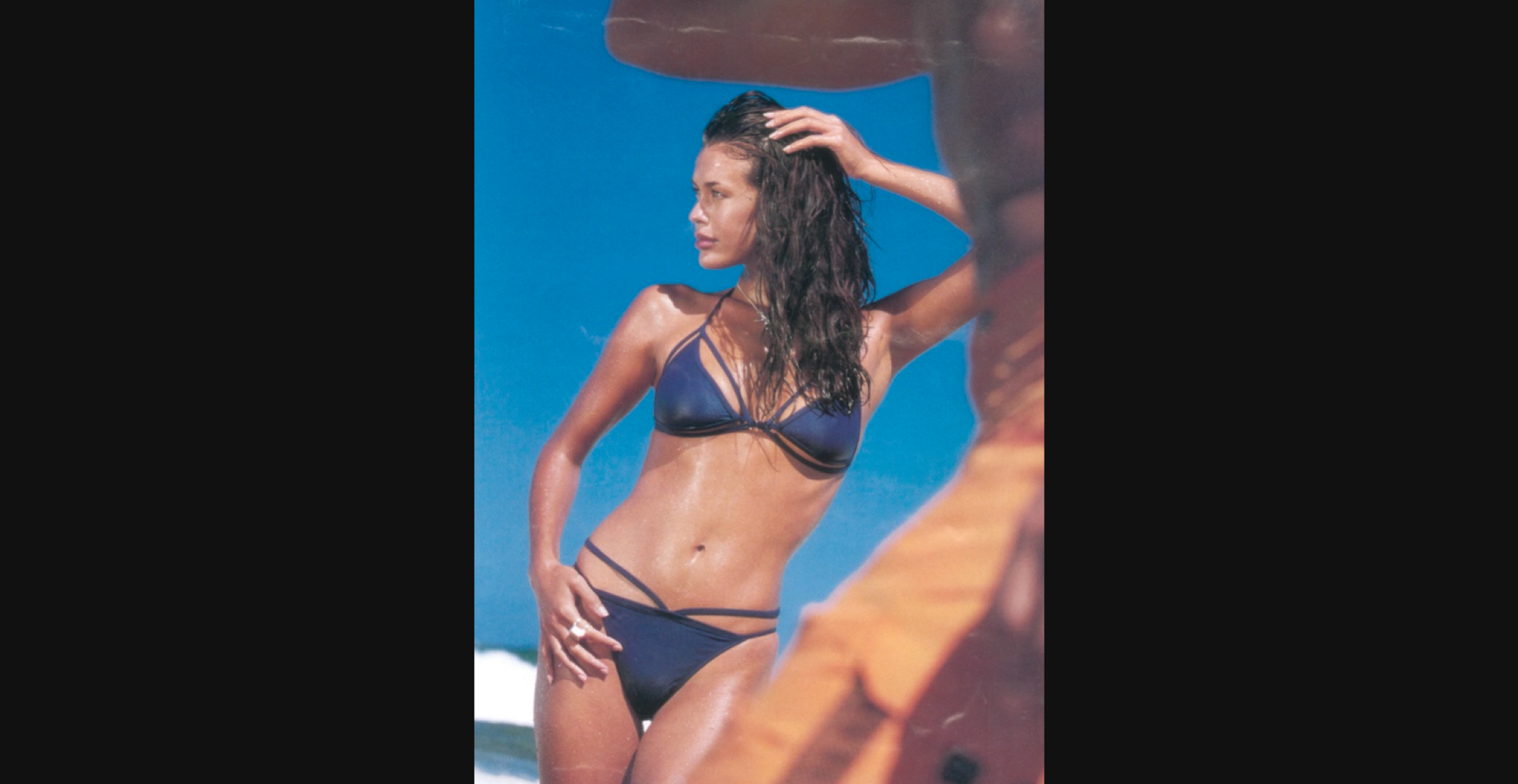 Colour photo of a model posing on a sunny beach in a dark blue strapped bikini, her wet hair pulled back with one hand against a bright blue sky backdrop