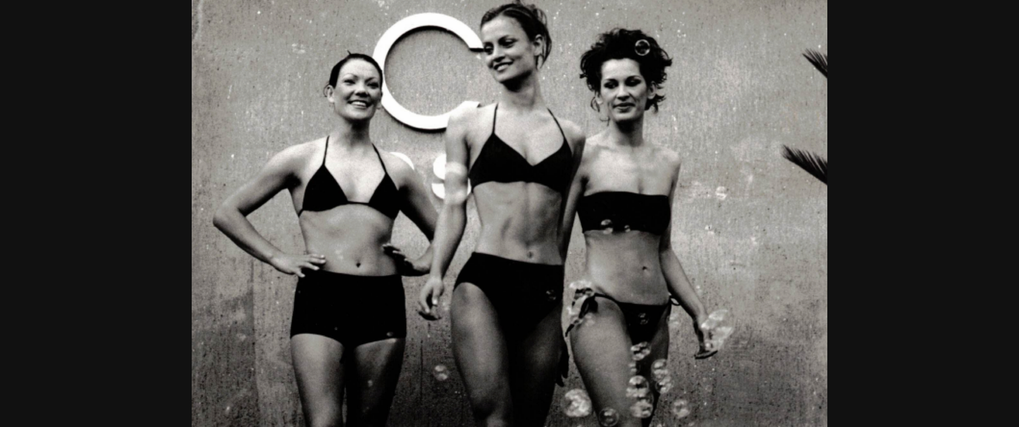 Black-and-white photo of three smiling models in black bikinis standing in front of a wall with a large “C” letter behind them.