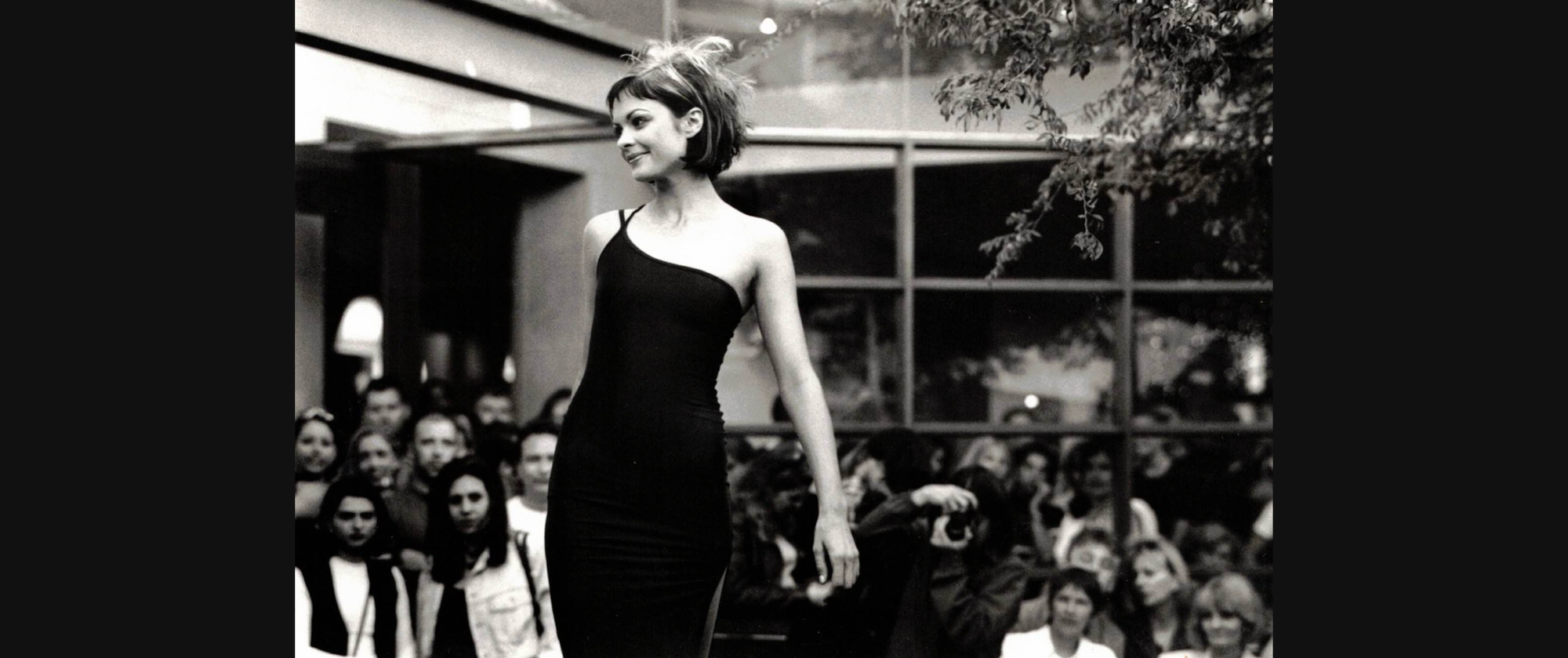 Black-and-white photo of a model wearing a one-shoulder, form-fitting black dress while walking past a crowd of onlookers.