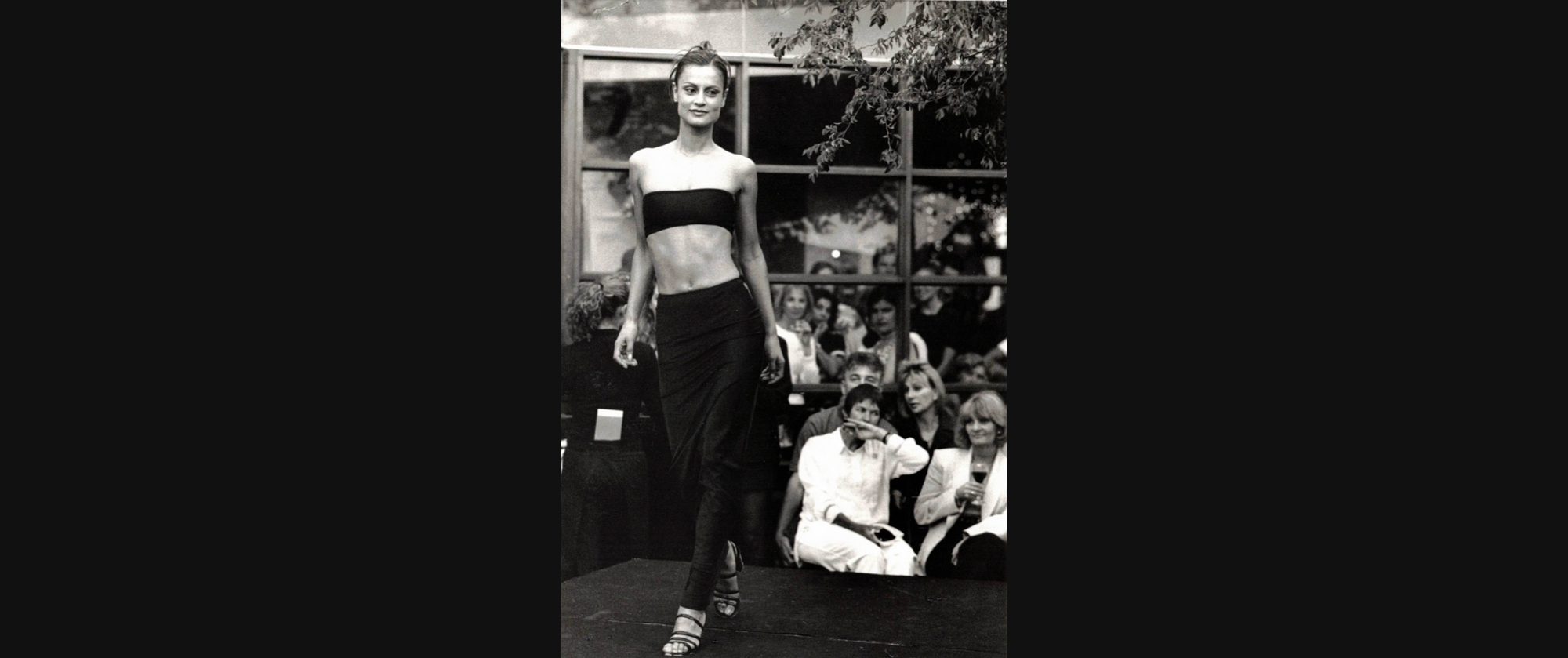 Black-and-white photo of a model in a strapless black bandeau top and a long black skirt walking on a runway, with an audience seated in the background