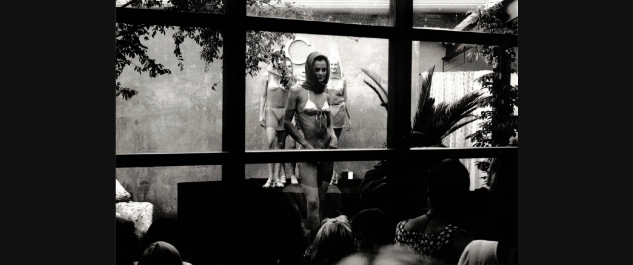 Black-and-white photo of several models in minimal swimwear and headscarves standing on a platform behind a large window, viewed by an audience in silhouette in the foreground