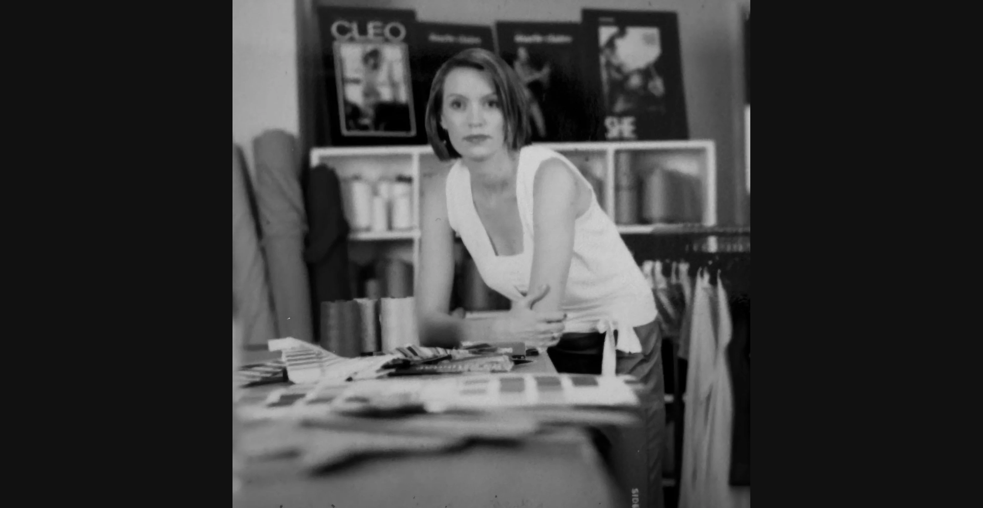 Black-and-white photo of a woman in a sleeveless top leaning on a table with fabric samples in a design studio, looking at the camera. Shelves in the background hold magazines and bolts of fabric